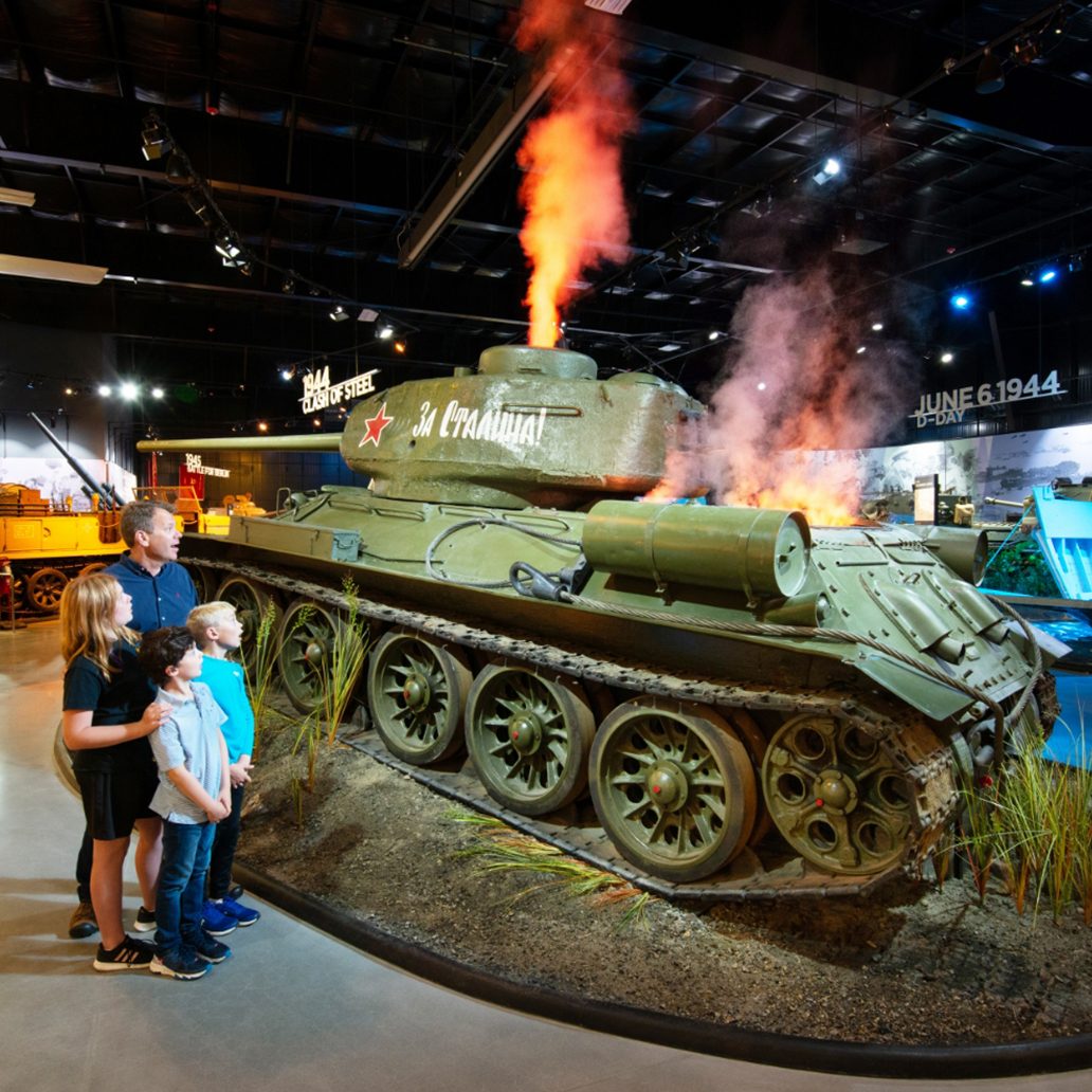 A family looks up at a restored Soviet T-34 tank amid smoke effects in the American Heritage Museum WWII exhibit hall